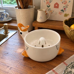 Pode incluir: Um espremedor de citrinos em cerâmica branca com formato de ganso, apresentando bico laranja, pés laranja e um olho preto. A taça tem um cone central com orifícios de drenagem e o exterior é decorado com desenhos de linhas pretas simples.