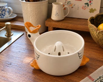 May include: A white ceramic citrus juicer shaped like a goose, featuring an orange beak, orange feet, and a black eye. The bowl has a central reamer with drainage holes, and the exterior is decorated with simple black line drawings of wings.
