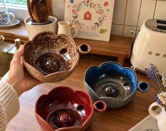 May include: Three ceramic berry bowls shaped like flowers with scalloped edges. Each bowl features a central perforated dome, a side handle, and a glossy glaze in speckled brown, deep red, or blue and grey. The bowls are designed for washing fruit.
