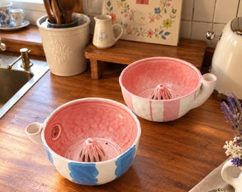 May include: Two ceramic citrus juicers with a central reamer and a side spout. One juicer features a pink interior and white exterior with pink stripes, while the other has a pink interior and white exterior with blue stripes.