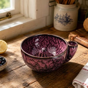 May include: A ceramic citrus juicer featuring a deep purple and black mottled glaze. The bowl-shaped vessel includes a central reamer for extracting juice and a small side handle for gripping, finished with a smooth, glossy texture.