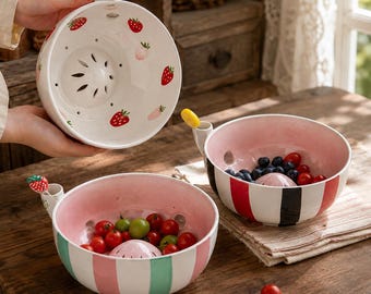 May include: Three ceramic colander bowls with handles. One bowl is white with red strawberry patterns. Another bowl features pink and green vertical stripes with a strawberry topper. The third bowl has pink and black vertical stripes with a yellow topper.