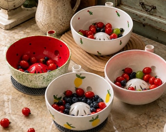 May include: Three ceramic berry bowls with integrated colander centers. One bowl is red with a speckled green exterior, one is white with green tree motifs, and one is white with yellow lemon patterns, all containing fresh berries and tomatoes.
