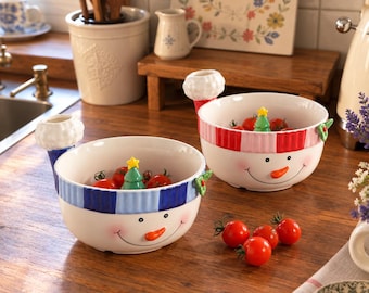 May include: Two ceramic snowman bowls featuring a white base, a smiling face with an orange carrot nose, and a striped rim. One bowl has a blue striped rim, the other a red striped rim. Each bowl includes a small green Christmas tree accent.