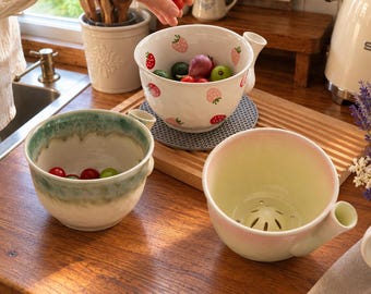 May include: Three ceramic berry colander bowls with side handles. One bowl features a white base with red and pink strawberry illustrations. Another bowl has a green and white ombre glaze. The third bowl is a pale pink and cream color with a drainage base.