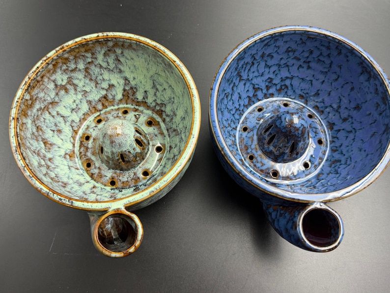 May include: Two ceramic citrus juicers with a speckled glaze finish. One juicer is light green with brown accents, and the other is blue with dark blue speckles. Both feature a central reamer with drainage holes and a side spout.