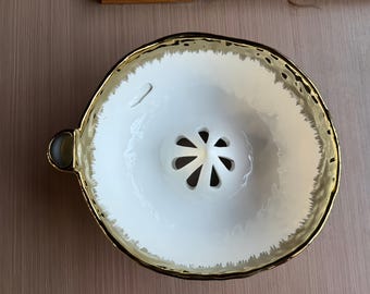 May include: A white ceramic citrus juicer featuring a central reamer with eight drainage slots. The bowl has a textured, uneven rim finished with a metallic gold glaze that fades into the white interior. A small handle extends from the side.