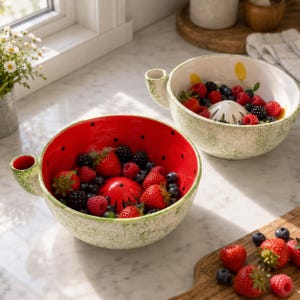 Pode incluir: Dois recipientes de cerâmica para frutos silvestres com exterior verde salpicado e interior vermelho brilhante, apresentando um cone de drenagem central e uma pega lateral. Os recipientes estão cheios de morangos, mirtilos e amoras frescas.