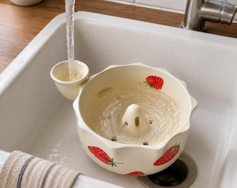 May include: A cream-colored ceramic berry colander featuring a scalloped rim and a painted strawberry pattern. The bowl has a central drainage cone and a side spout positioned under a running kitchen faucet, designed for washing fresh produce.