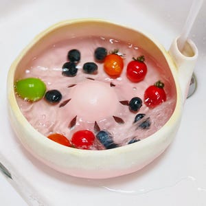 Peut inclure: Une passoire à baies ronde en céramique rose clair avec un dôme central surélevé et des fentes de drainage. Le bol est rempli d'eau, de myrtilles fraîches, de tomates cerises rouges et d'un raisin vert, avec un filet d'eau coulant sur le côté.