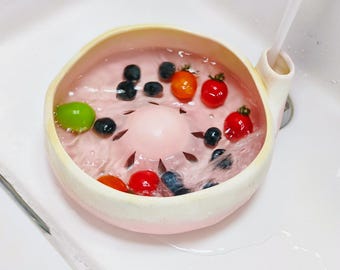 May include: A round, light pink ceramic berry colander with a central raised dome and drainage slits. The bowl is filled with water and fresh blueberries, red cherry tomatoes, and a single green grape, with a stream of water flowing into the side.