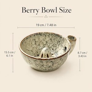 May include: A ceramic berry bowl with a speckled cream and brown glaze, featuring a central cone and a side handle. Text reads Berry Bowl Size. Dimensions are 7.48 inches wide, 6.1 inches deep, and 3.43 inches high, as indicated by the labels.