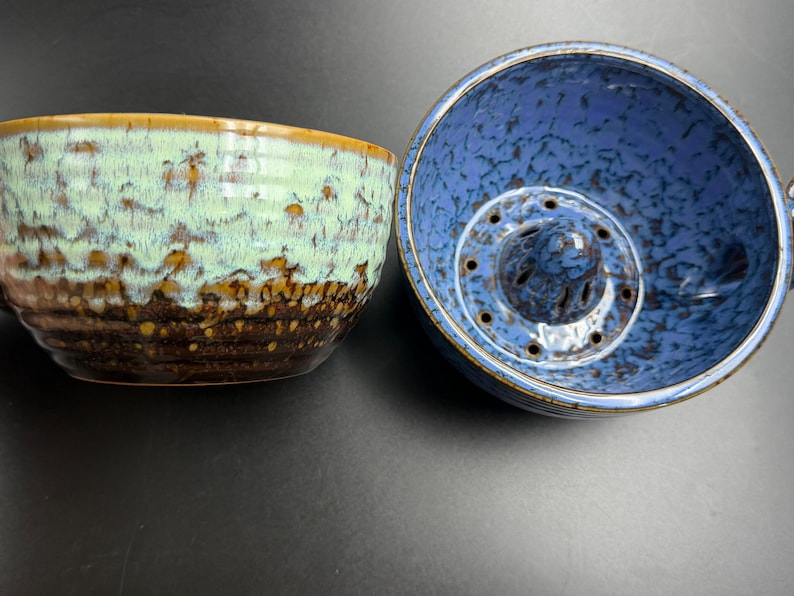 May include: Two ceramic bowls with a speckled glaze. The bowl on the left features a light mint green upper section transitioning into a dark brown base. The bowl on the right is blue with a central raised dome containing small drainage holes.
