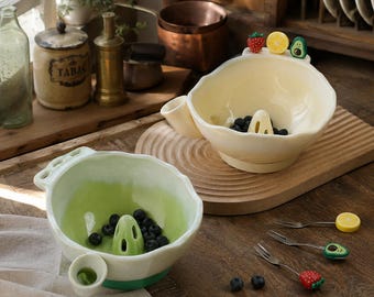 May include: Two ceramic berry bowls with central drainage cones and side pouring spouts. One bowl is cream-colored with strawberry, lemon, and avocado accents, while the other is light green. Three small fruit-topped metal forks are included.