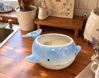 May include: A ceramic whale-shaped bowl with a glossy finish, featuring a gradient of light blue fading into white. The bowl has small black eyes, a simple curved mouth, two side fins, and a tail fin that extends upward from the back rim.