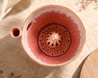 May include: A round ceramic berry bowl with a glossy pink and cream glaze. The interior features a raised central drainage dome surrounded by a ring of small circular holes for rinsing fruit, and a small side handle for easy gripping.