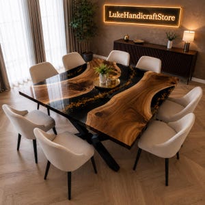 May include: A square dining table featuring a live edge wood slab with a glossy black resin river inlay containing gold flakes, supported by a black metal base, surrounded by eight upholstered cream-colored chairs with dark legs.