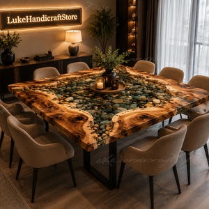 Könnte beinhalten: Ein quadratischer Esstisch mit einer Naturholzkante, die eine mittige Einlage aus glatten, mehrfarbigen Flusskieselsteinen in klarem Harz umschließt. Der Tisch steht auf schwarzen Metallbeinen und ist von acht beigefarbenen Polsterstühlen umgeben.