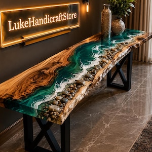 Può includere: Un lungo tavolo console caratterizzato da un piano in legno naturale con bordo vivo e un design a fiume centrale realizzato in resina color ottanio, accenti di schiuma bianca e ciottoli incastonati. Il tavolo è sostenuto da gambe in metallo nero a forma di X sotto un'insegna con la scritta LukeHandicraftStore.