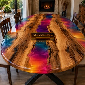 Op de afbeelding: Een ovale eettafel met een natuurlijk houtnerfoppervlak en een centraal rivierontwerp van levendig roze, oranje en blauwe hars. Een klein rechthoekig bordje met de tekst LukeHandicraftStore staat in het midden van de tafel.