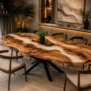 Puede incluir: Una mesa de comedor ovalada con un tablero de madera natural de borde irregular y una incrustación central de resina blanca. La mesa se apoya sobre una base de metal negro y está rodeada por seis sillas de madera con cojines claros.