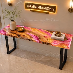 Puede incluir: Una mesa consola rectangular con un tablero de madera de borde natural con remolinos de resina rosa y naranja vibrantes, sostenida por dos patas de metal negro en forma de U. Un jarrón de cerámica, un cuenco dorado y un libro decoran la superficie.