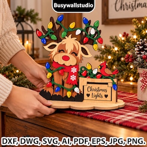 Può includere: Una decorazione in legno intagliato raffigurante una mucca delle Highland con una sciarpa rossa, avvolta in luci natalizie colorate. Un piccolo cardinale rosso è posato su un cartello con la scritta Christmas Lights e due cuori.