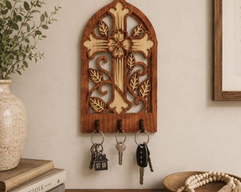 May include: A wall-mounted wooden key holder featuring an arched frame with a cutout cross and floral design. The piece has three dark hooks at the bottom, each holding a set of keys. The wood displays a two-tone finish with light and dark brown sections.