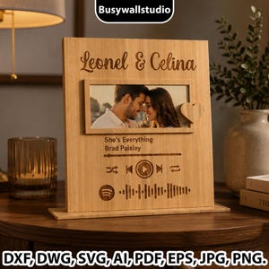 May include: A rectangular light wood plaque featuring the engraved names Leonel & Celina above a central photo frame containing a picture of a couple. Below the photo, the text She's Everything by Brad Paisley appears with a music player interface, a play button, and a Spotify-style sound wave graphic. A small wooden heart is attached to the right side of the frame.