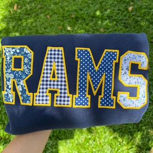 May include: A navy blue sweatshirt featuring the word RAMS in large block letters. Each letter is outlined in yellow and filled with a different pattern: blue floral, blue and white gingham, blue with white polka dots, and light blue floral.
