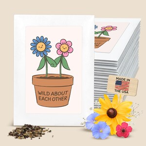 May include: A stack of white greeting cards featuring a cartoon illustration of a blue flower and a pink flower in a brown pot with the text WILD ABOUT EACH OTHER. A small wooden sign with a US flag and the text MADE IN THE USA is included.