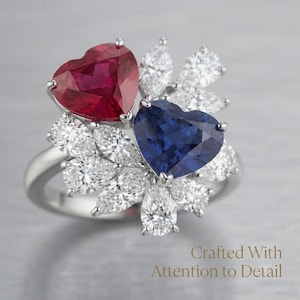 May include: A silver-toned ring featuring a large heart-shaped red gemstone and a large heart-shaped blue gemstone, surrounded by multiple pear-shaped clear stones. The text "Crafted With Attention to Detail" is visible in the lower right corner.