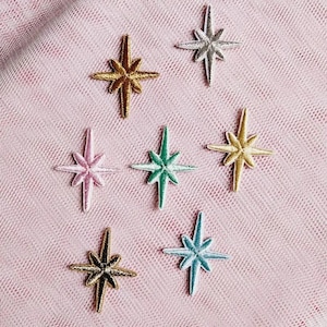 Könnte beinhalten: Sieben gestickte sternförmige Aufnäher in verschiedenen Farben, darunter Gold, Silber, Rosa, Grün und Hellblau. Jeder Aufnäher zeigt ein mehrzackiges Sterndesign mit gestickten Details, die in einem Streumuster auf einem strukturierten rosa Stoff angeordnet sind.
