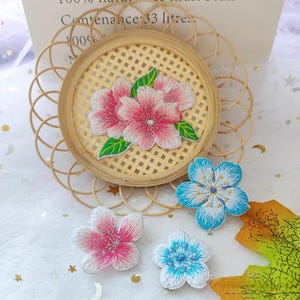 Può includere: Una collezione di toppe ricamate a forma di fiore con un gruppo di fiori rosa e foglie verdi su un vassoio rotondo di bambù, accompagnate da due toppe individuali a cinque petali nelle tonalità del rosa e del blu con dettagli bianchi.