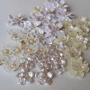 Puede incluir: Una colección de adornos florales decorativos con pétalos iridiscentes en tonos blanco y crema. Algunas flores están hechas de acrílico en capas con centros de cuentas, mientras que otras son arreglos intrincados de pequeñas perlas blancas.