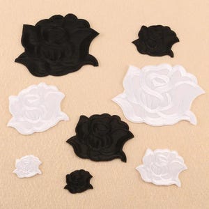 May include: A collection of eight embroidered rose patches in varying sizes, featuring four solid black roses and four solid white roses. Each patch displays detailed stitching that forms the layered petals of a blooming rose flower design.