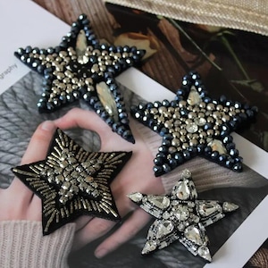 May include: Four star-shaped patches featuring intricate beadwork and crystal embellishments. Two stars are covered in dark metallic beads and clear crystals, one features silver tube beads on black felt, and one has clear faceted crystals.