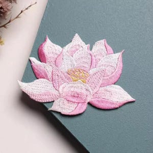 May include: An embroidered patch featuring a blooming lotus flower with layered petals in shades of soft pink and white. The center of the flower displays a textured yellow seed pod with small circular details, all rendered in dense thread work.