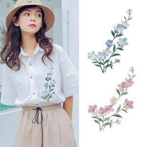 May include: A white short-sleeved button-down shirt featuring an embroidered blue floral sprig on the chest. Two separate floral embroidery patches are shown next to the shirt, one with blue flowers and one with pink flowers, each on green stems.