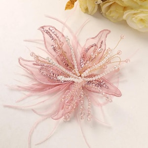 May include: A decorative floral accessory in soft pink, featuring sheer fabric petals embellished with shimmering sequins and beads. The center is adorned with clusters of white pearls and delicate pink feathers extending outward from the base.
