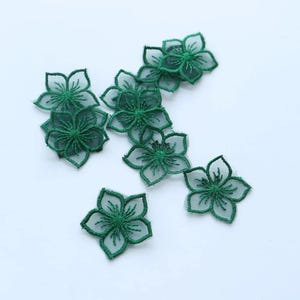 Puede incluir: Un conjunto de siete aplicaciones florales de color verde oscuro, cada una con un diseño de flor de cinco pétalos hecho de tela de malla fina y transparente con bordes bordados intrincados y costuras detalladas que irradian desde el centro.