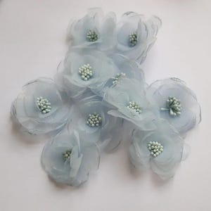 Puede incluir: Un conjunto de ocho flores de tela azul claro con pétalos superpuestos y translúcidos. Cada flor presenta un grupo de pequeñas cuentas de color verde pálido en el centro, creando una apariencia texturizada. Las flores están dispuestas en grupo.