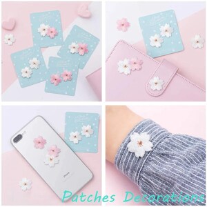 May include: A collection of small, embroidered cherry blossom patches in shades of white and soft pink. These floral appliques are displayed on light blue backing cards, attached to a light pink notebook, a smartphone, and a striped shirt sleeve.