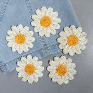 Puede incluir: Cuatro parches de flores tipo margarita en blanco y amarillo dispuestos sobre un fondo de tela vaquera azul claro. Cada parche presenta un centro amarillo circular rodeado de pétalos blancos, con un diseño de hilo texturizado.