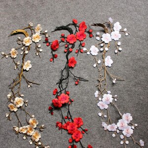 May include: Three embroidered fabric appliques featuring flowering branches. One branch is gold with cream flowers, one is black with vibrant red flowers, and one is brown with white and pale pink flowers. Each piece shows detailed floral stems.