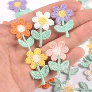 Puede incluir: Una colección de pequeños parches bordados de flores en varios colores, incluyendo naranja, blanco, morado, amarillo y rosa. Cada parche presenta una flor con un centro contrastante, un tallo delgado y dos pequeñas hojas de color verde menta.