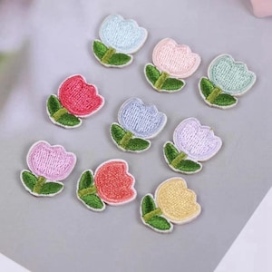 May include: A collection of nine small embroidered tulip patches in various pastel and vibrant colours including pink, blue, yellow, and lavender. Each patch features a rounded flower head with a green stem and two leaves, finished with a white border.