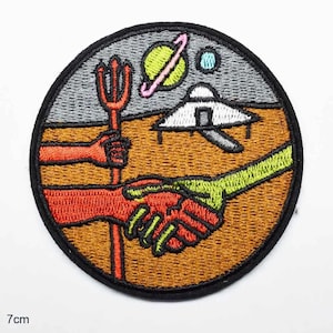 May include: A circular embroidered patch measuring 7 cm in diameter, featuring a handshake between a red hand and a green hand. The scene includes a red trident, a white UFO, a yellow planet with a pink ring, and a small blue planet on a brown surface.