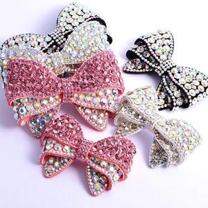 Op de afbeelding: Een verzameling van vier strikvormige haaraccessoires versierd met sprankelende strass-steentjes. Twee strikken zijn voorzien van iriserende heldere kristallen, terwijl twee strikken roze kristallen combineren met iriserende accenten.