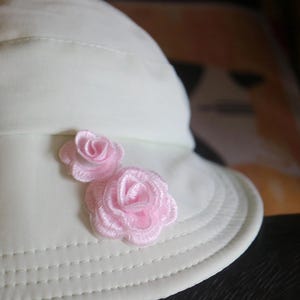 Puede incluir: Un sombrero de pescador color crema con dos aplicaciones de rosas bordadas en color rosa claro en el lateral. El sombrero tiene un ala ancha con costuras y una estructura de tela lisa de color sólido con detalles de hilo visibles en el borde.